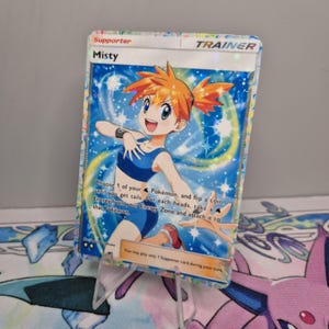 May include: A holographic "Misty" Trainer card from the Pokémon trading card game. The card features a cartoon illustration of a girl with orange hair and blue clothing. The card has text that describes the card's function and is displayed on a clear stand.