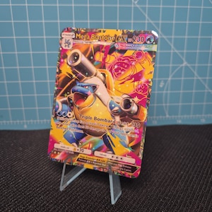 Peut inclure: Carte Pokémon Mega Blastoise EX avec un personnage bleu et jaune avec des canons. Le texte "Triple Bombardment 130" et "HP 230" est visible. Le fond est un mélange d'orange, de rose et de jaune.