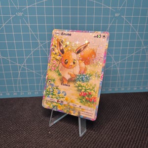 Op de afbeelding: Een holografische Eevee Pokémon-verzamelkaart op een heldere acryl standaard. De kaart toont Eevee in een bloemenomgeving, met de tekst "Eevee" en "HP60". De achtergrond is een blauw raster.