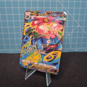 Puede incluir: Una carta Pokémon Mega Venusaur EX, con un personaje azul y rosa con diseños florales. Se puede ver el texto "Critical Bloom" y "HP 240". La carta se muestra en un soporte de plástico transparente.