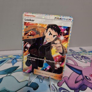 May include: A holographic "Looker" Trainer card from the Pokémon Trading Card Game. The card features an illustration of a man in a suit pointing forward, with text that reads "Looker" and "Trainer". The card is supported by a clear plastic stand.