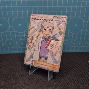 Könnte beinhalten: Eine holografische Trainerkarte mit dem Titel "Professor's Research" mit einer Illustration von Professor Eich. Die Karte hat einen weißen Rand und den Text "Draw 2 cards". Die Karte wird auf einem transparenten Kunststoffständer präsentiert.