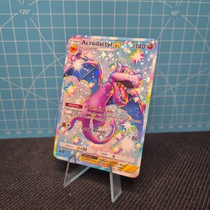 May include: A holographic Acrodactyl EX Pokémon card, featuring a purple and blue dragon-like creature with text that reads "Primeval Law" and "Land Crush". The card is displayed on a clear acrylic stand.