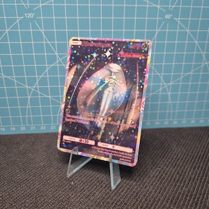 May include: A holographic Pheromosa Pokémon trading card, displayed on a clear acrylic stand. The card features a detailed illustration of Pheromosa with the text "Jump Blues" and "20". The card has a colorful, glittery background.