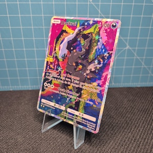 Op de afbeelding: Een holografische Darkrai-verzamelkaart met een levendige, veelkleurige illustratie van het personage. De kaart toont de naam Darkrai, 100 HP, de vaardigheid Bad Dreams en de aanval Dark Slumber. Het staat rechtop in een doorzichtige standaard.