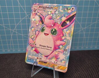 Könnte beinhalten: Eine holografische Wigglytuff EX Pokémon-Sammelkarte, präsentiert auf einem transparenten Acrylständer. Die Karte zeigt einen rosa, runden Charakter mit großen Augen und dem Text "Sleepy Song". Die Karte hat 140 HP und einen Sternenhintergrund.