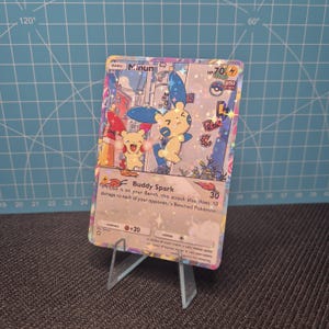 May include: A holographic Minun Pokémon trading card, featuring two cartoon characters with blue and yellow accents. The card is displayed on a clear plastic stand against a blue grid background. Text includes "Buddy Spark" and "HP 70".
