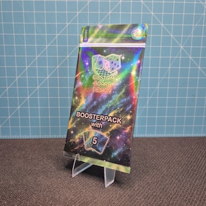 Könnte beinhalten: Eine rechteckige Booster-Packung mit einem holografischen Hintergrund im Galaxie-Design mit Sternen und Nebelwolken. Der Text lautet Custom Board Design, Boosterpack with 5 und CBD01. Oben ist eine Grafik eines Würfels und Spielkarten zu sehen.