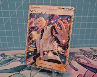 Puede incluir: Una carta de Entrenador "Cyrus" holográfica del Juego de Cartas Coleccionables Pokémon. La carta presenta un personaje con cabello azul y un atuendo blanco y negro. El texto de la carta incluye "Supporter" e instrucciones del juego. El fondo es una galaxia colorida.