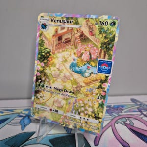 Könnte beinhalten: Eine Venusaur Pokémon-Sammelkarte mit einer farbenfrohen Illustration eines Hauses und Gartens. Die Karte zeigt den Text "Venusaur", "HP 160" und "Mega Drain". Die Karte wird auf einem transparenten Ständer präsentiert.