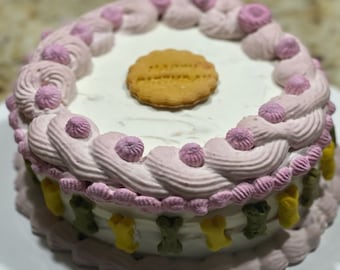 May include: A small, round cake with white frosting and pink frosting swirls. The cake is decorated with small, colorful bone-shaped candies around the base and a round biscuit on top. The cake is on a white plate.