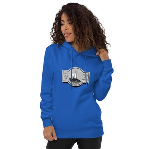 Puede incluir: Una sudadera con capucha azul real con un gráfico de un faro en una isla rocosa con el texto "Cabot Cove, ME" y "Jessica B Fletcher". La sudadera con capucha tiene una capucha con cordón y un bolsillo de canguro.