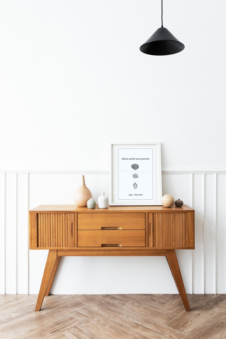 May include: A white framed print with the text "Salt air, and the rust on your door,  still smells like home."  The print is sitting on a wooden console table with two drawers. There are three decorative vases on the table.