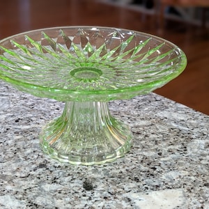 May include: A green glass cake stand with a round, diamond-patterned top and a pedestal base. The stand is made of clear glass with a slight green tint, and it sits on a speckled countertop.