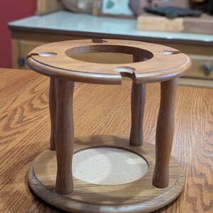 May include: A wooden wine glass holder with a circular top and base, connected by four curved legs. The top has cutouts for glasses, and the base has a cork insert. The holder is made of light brown wood and sits on a wooden table.
