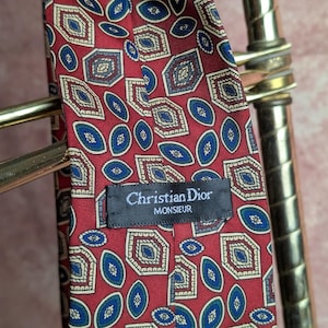 May include: A red necktie featuring a repeating pattern of blue oval shapes and geometric hexagonal motifs. A black fabric label is stitched onto the center, displaying the white text Christian Dior MONSIEUR in a clear, legible font.