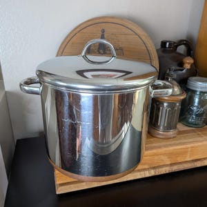 May include: A stainless steel cooking pot with a lid, featuring a silver handle and side handles. The pot has a shiny, reflective surface. The pot is sitting on a wooden surface, with other kitchen items in the background.