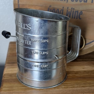 May include: A vintage metal flour sifter with a handle and a black knob. The sifter has measurements for one, two, and three cups. The text "WELL'S" and "SIFTER" are visible. The background includes a wooden sign with the text "Good Friends, Good Wine."