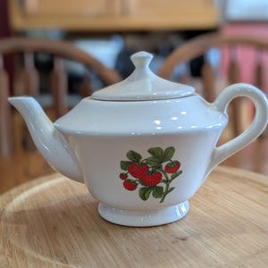 May include: A white ceramic teapot with a lid and a curved handle. The teapot features a strawberry design with red berries and green leaves. The teapot is sitting on a wooden tray.