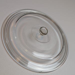 May include: A circular, clear glass lid featuring a smooth, raised central knob handle. The lid has a stepped, concentric rim design with a subtle, fine-textured pattern visible on the interior surface of the glass, providing a clear view.