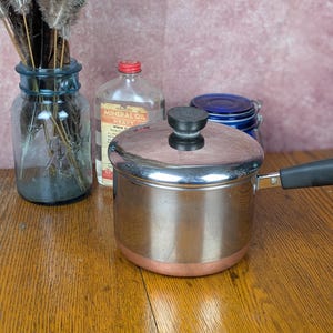 May include: A stainless steel saucepan with a copper-colored base, a matching lid with a black knob, and a black handle. The pot features a polished metallic finish and a sturdy construction, designed for stovetop use in a kitchen environment.