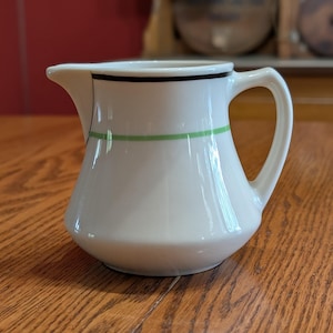 May include: A white ceramic creamer pitcher featuring a smooth, rounded body, a small pouring spout, and a curved handle. The exterior is decorated with a thin black horizontal line near the rim and a wider, vibrant green horizontal band below it.