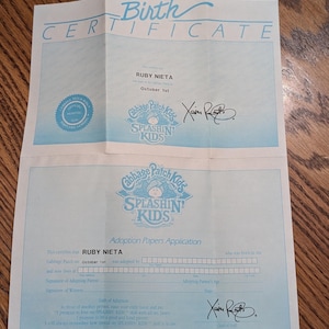 May include: A light blue Cabbage Patch Kids Splashin Kids birth certificate and adoption papers application document. The paper features the name Ruby Nieta, the date October 1st, the official hospital seal, and the signature of Xavier Roberts.