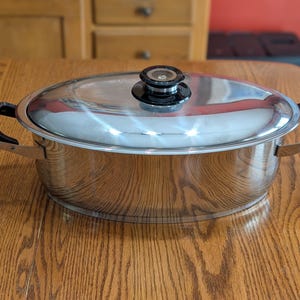 May include: An oval stainless steel roasting pan with a polished finish, featuring two black side handles and a matching stainless steel lid with a black knob handle. The cookware has a wide, shallow body and a reflective surface throughout.