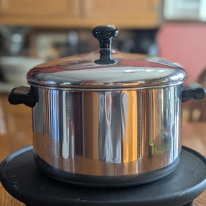 May include: A stainless steel pot with a lid and black handles. The pot is sitting on a black, round, electric hot plate. The background is a blurred kitchen scene with wooden cabinets and a table.