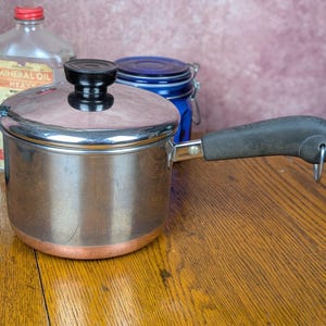 May include: A stainless steel saucepan with a copper base, featuring a matching lid with a black knob. The pan has a long, dark gray handle with a metal hanging loop at the end. The exterior surface is reflective, showing a cylindrical shape.