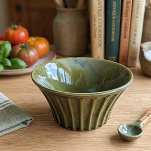 May include: A green ceramic bowl with a fluted design, a small olive wood spoon, and a folded green and white striped linen napkin sit on a wooden table. Tomatoes and basil are on a plate in the background.
