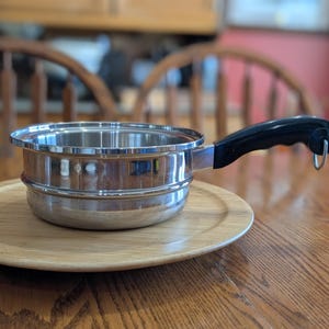 May include: A stainless steel saucepan with a black handle and a silver rim. The pan sits on a round wooden tray. The handle has a loop for hanging. The pan is on a wooden table.