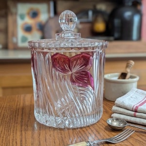 May include: A clear glass jar with a lid, featuring a decorative floral design in shades of pink. The jar has a textured, swirled pattern and sits on a wooden surface. A spoon, fork, and knife are also visible.
