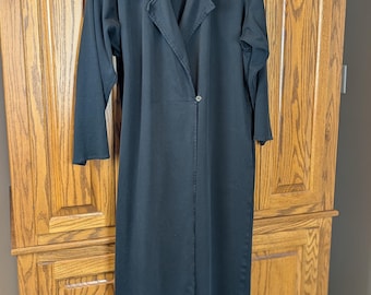 May include: A long, black dress with long sleeves and a collared neckline. The dress has a single button closure and a tailored fit. The garment is hanging on a hanger against a wooden cabinet.