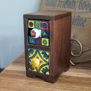 May include: A small, wooden two-drawer cabinet with colorful, patterned ceramic drawer fronts. The drawers feature floral and geometric designs in blue, yellow, red, and green. The knobs are white and yellow. The cabinet is on a wooden surface.