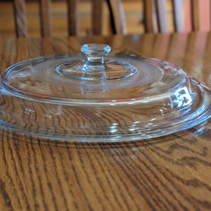 May include: A clear glass lid with a small knob handle. The lid is circular with a slightly raised rim and a textured center. The lid is designed to fit on a round dish. The lid is sitting on a wooden table.