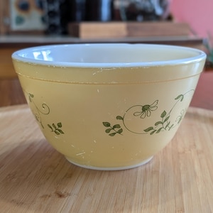May include: A vintage yellow mixing bowl with a white rim and green floral design. The bowl is set on a wooden tray. The bowl's exterior features a delicate pattern of green flowers and leaves.