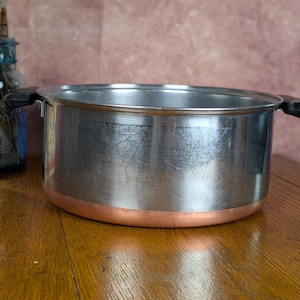 May include: A stainless steel cooking pot with a copper-colored base and two black side handles. The pot has a cylindrical shape with a polished metallic finish showing light surface scratches and a wide opening at the top for cooking tasks.