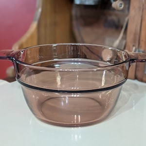 May include: A clear, round, glass bowl with two handles. The bowl has a dark brown rim and is made of a transparent, light brown glass. The bowl is designed for cooking and serving food.
