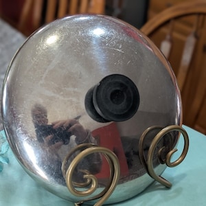 May include: A stainless steel pot lid with a black handle, resting on a decorative brass stand with a swirled design. The lid reflects the surrounding environment. Kitchenware.