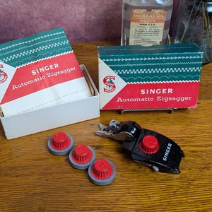 May include: A Singer Automatic Zigzagger sewing machine attachment with three red and gray cams, presented with its original white and red cardboard box featuring green decorative trim and the Singer logo, alongside a closed matching box.