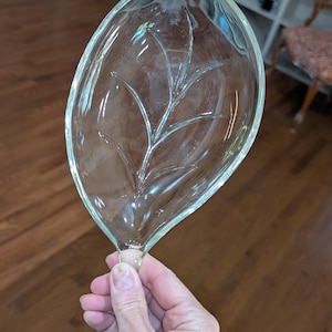 May include: Clear glass leaf-shaped dish with a detailed vein pattern. The dish is held by a hand, showcasing its delicate design. The leaf dish is a decorative item, suitable for serving or display.
