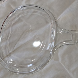 May include: A clear, oval-shaped glass lid with a handle. The lid is designed to fit a cooking dish. The handle is rectangular and extends from the side of the lid. The lid is resting on a white cloth.