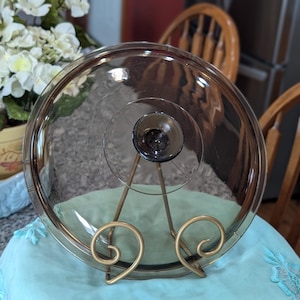 May include: A clear, round glass lid with a dark knob, resting on a gold-colored metal stand. The lid is slightly tinted and appears to be for a casserole dish. The stand has decorative swirls.