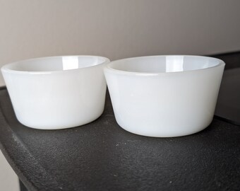 May include: Two small, white, round bowls with a slightly flared rim. The bowls are made of a solid, opaque material and sit on a dark surface. The bowls are empty and appear to be clean.