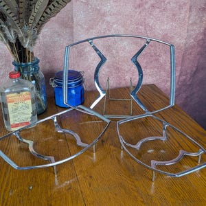 May include: Three chrome-plated metal wire racks with a rectangular frame and a wavy interior design. These stands feature a polished silver finish and are constructed with thin, sturdy metal rods, designed to hold items securely in place.
