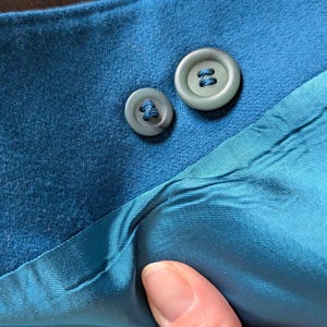 May include: Close-up of teal fabric with two matching buttons. The buttons are a muted teal color and have a four-hole design. The fabric appears to be a soft material, possibly suede or a similar texture. The lining is a lighter shade of teal.