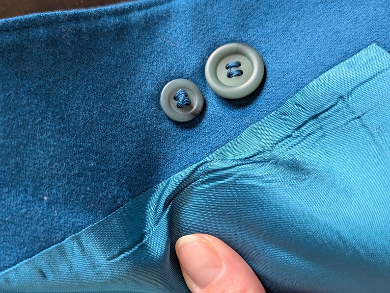 May include: Close-up of teal fabric with two matching buttons. The buttons are a muted teal color and have a four-hole design. The fabric appears to be a soft material, possibly suede or a similar texture. The lining is a lighter shade of teal.
