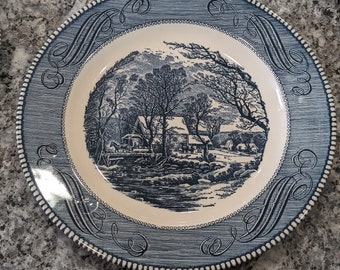 Puede incluir: Plato decorativo azul y blanco con un paisaje en el centro. El plato tiene un borde azul con volutas y un borde con cuentas. La imagen central representa una escena rural con árboles, una casa y un río. Diámetro aproximado de 25 cm.