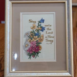 May include: A framed cross-stitch embroidery featuring a bouquet of yellow, blue, white, and pink flowers. The text reads, "Sing unto the Lord a New Song PS. 149:1" in brown lettering. The piece is enclosed in a gold-toned frame with a tan mat board.